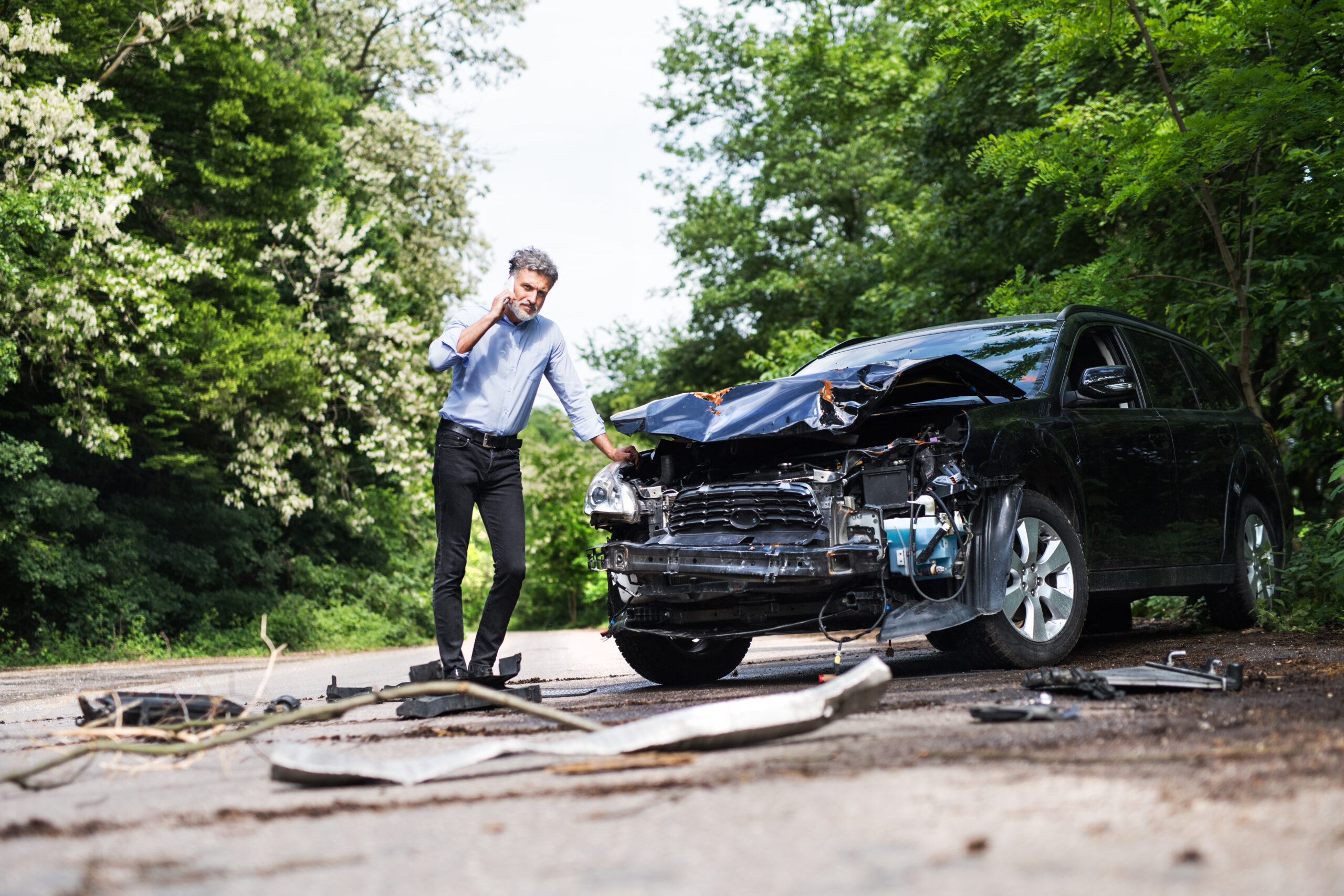 Man on phone standing beside a damaged car after a car accident on a rural road.