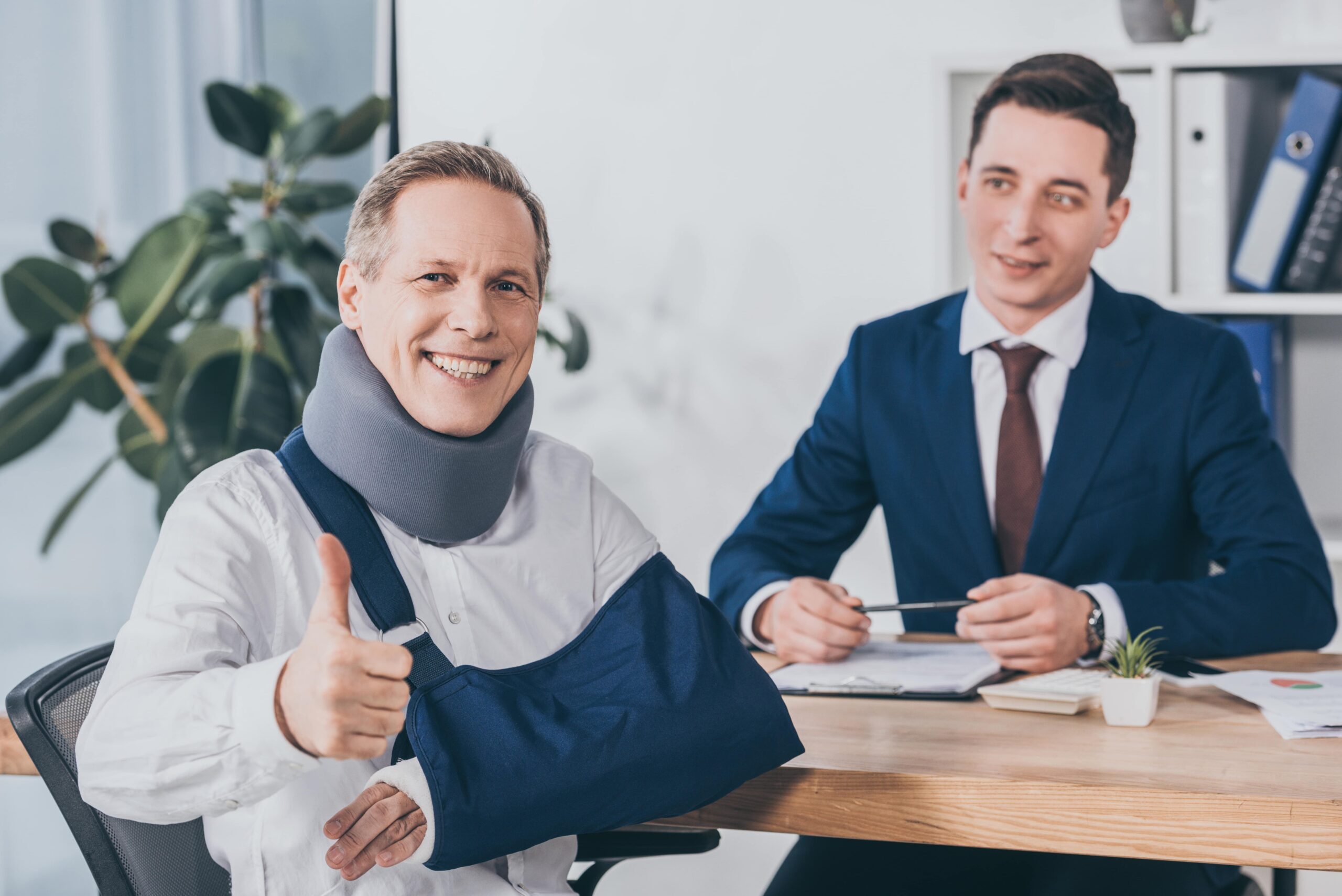 Injured man with neck brace and arm sling giving a thumbs up, sitting with a lawyer.